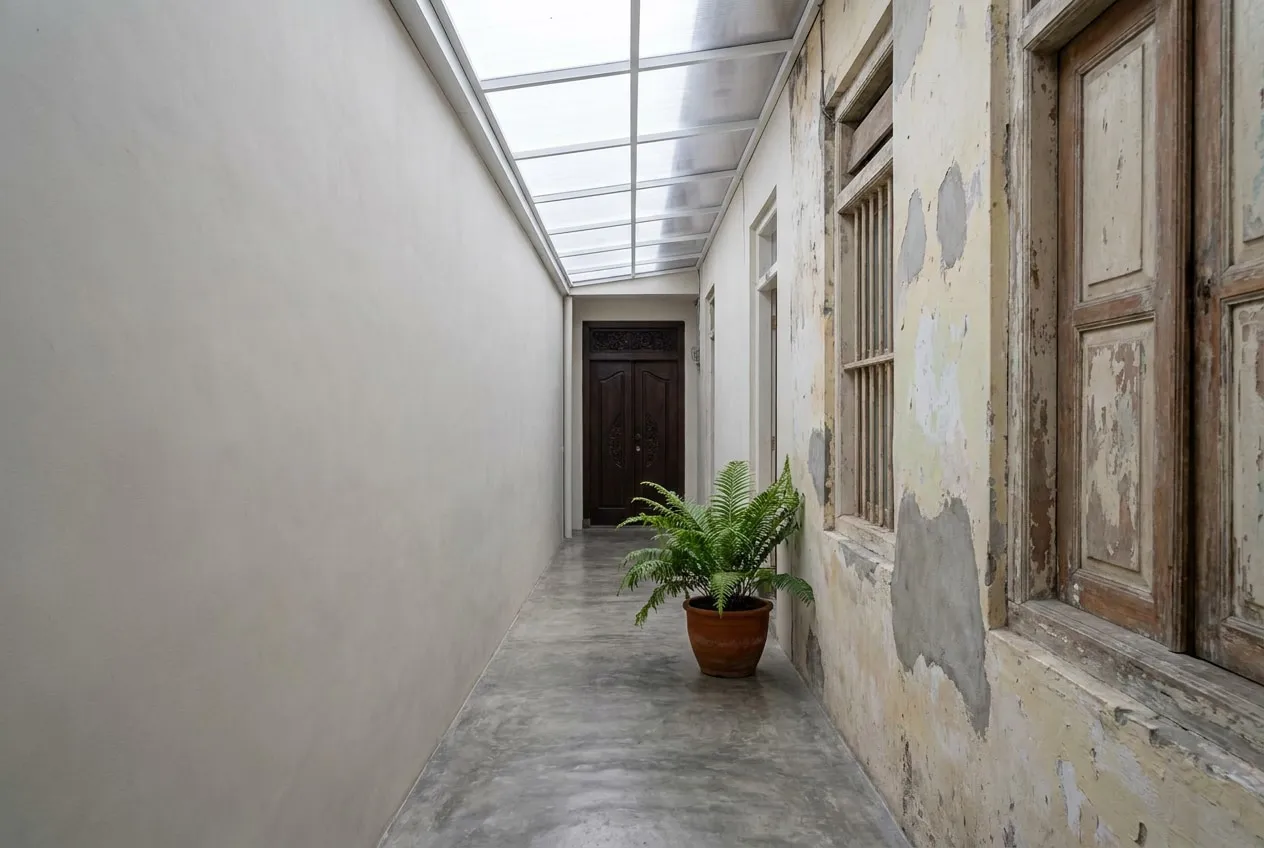 Lorong penghubung sempit beratap tembus cahaya, dengan dinding bata plester baru di satu sisi dan dinding lama berplester di sisi lain.