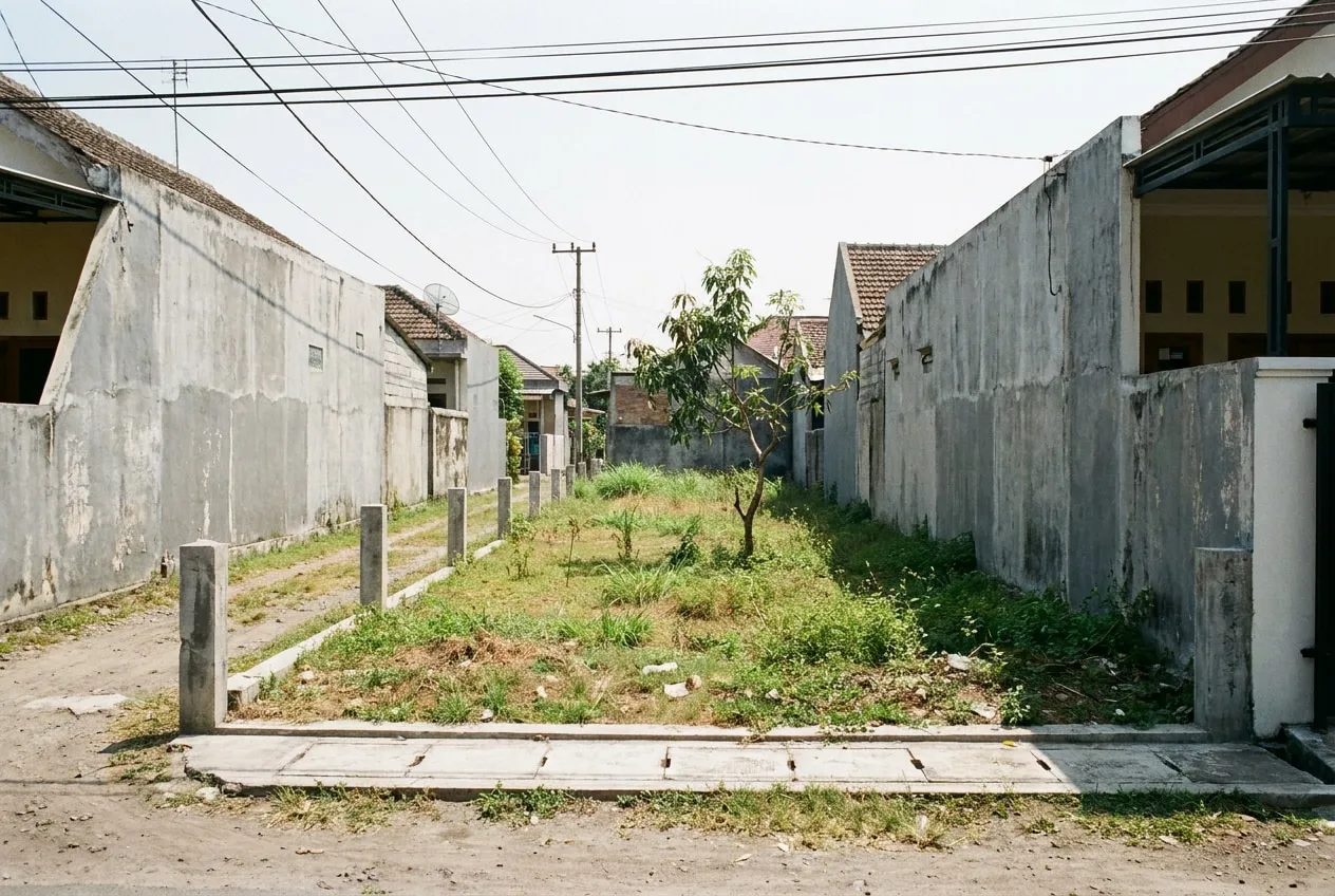 Lahan kosong memanjang dengan rumput dan satu pohon, dibatasi dinding rumah tetangga di kedua sisi, difoto dari jalan lingkungan pada siang hari.