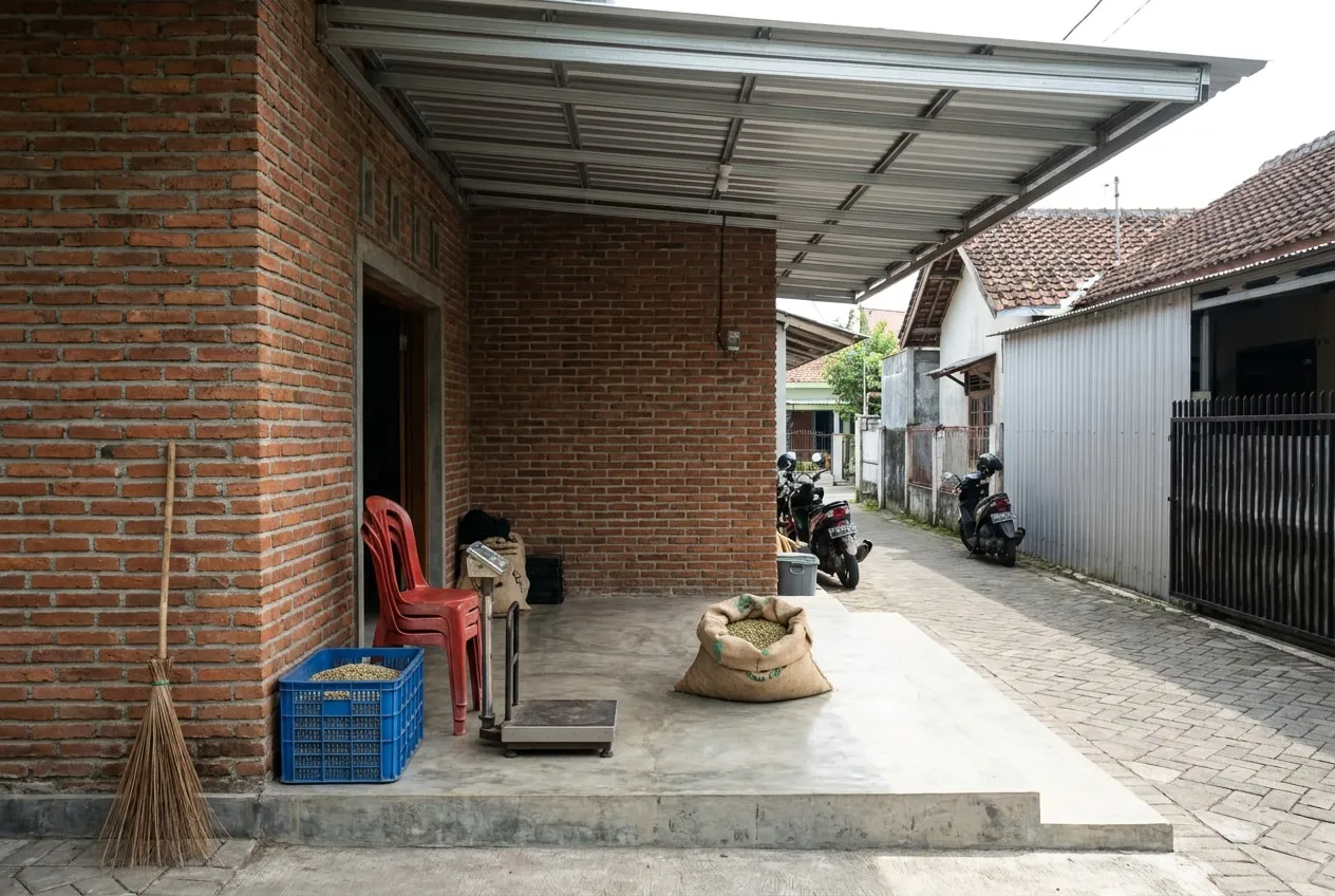 Teras beratap dangkal dengan karung kopi terbuka, timbangan, dan kursi plastik, menghadap gang lingkungan.